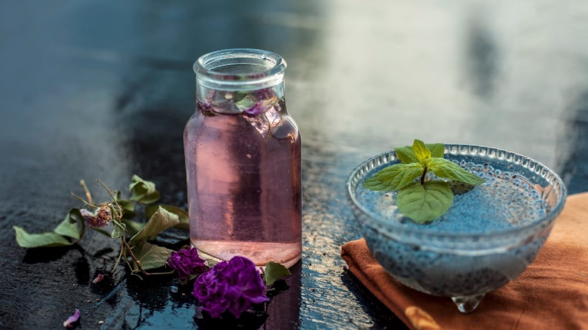 Ein Glas mit dem Gulab Sharbat auf einem dunklen Tisch mit einer blauen Glasschale und Blüten.