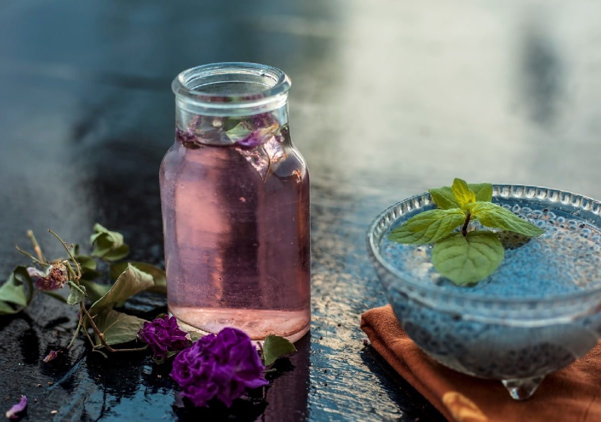 Ein Glas mit dem Gulab Sharbat auf einem dunklen Tisch mit einer blauen Glasschale und Blüten.