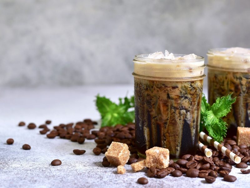 Zwei Gläser griechischer Frappé, drumherum Kaffeebohnen, Zuckerwürfel und Strohhalme.