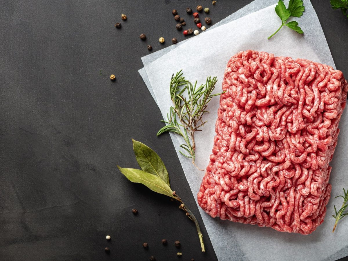 aufgetautes Hackfleisch liegt auf einem dunklen Untergrund. Daneben liegen frische Kräuter und Pfefferkörner.