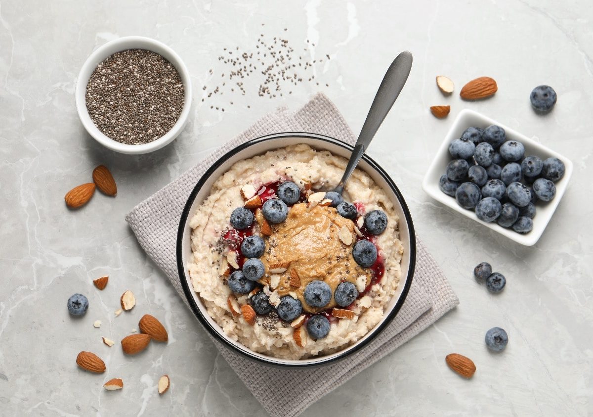 Eine Schale mit dem Mandel-Porridge auf einem hellgrauen Tisch. Um die Schale herum sind Mandeln, Blaubeeren und Chiasamen.