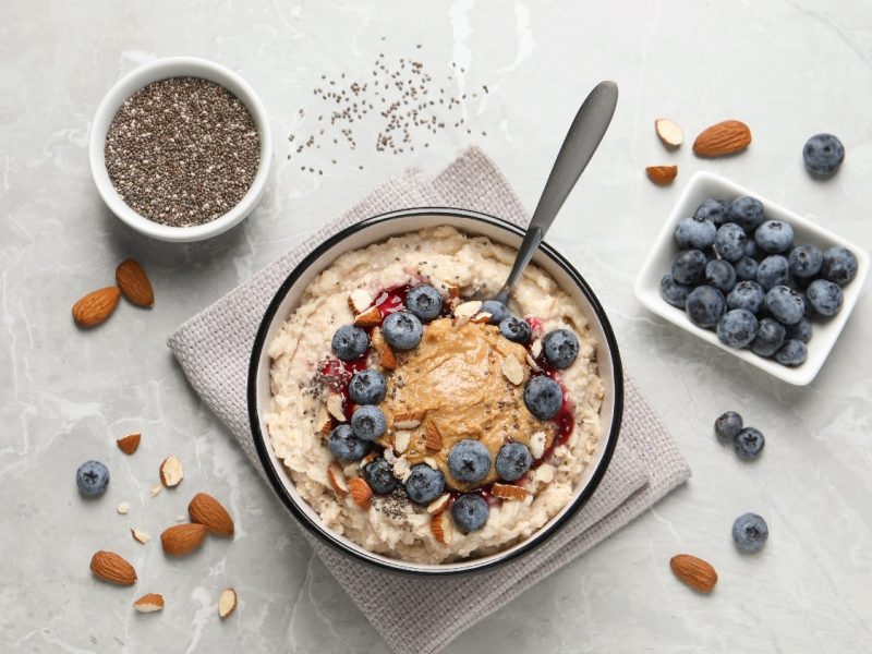 Eine Schale mit dem Mandel-Porridge auf einem hellgrauen Tisch. Um die Schale herum sind Mandeln, Blaubeeren und Chiasamen.