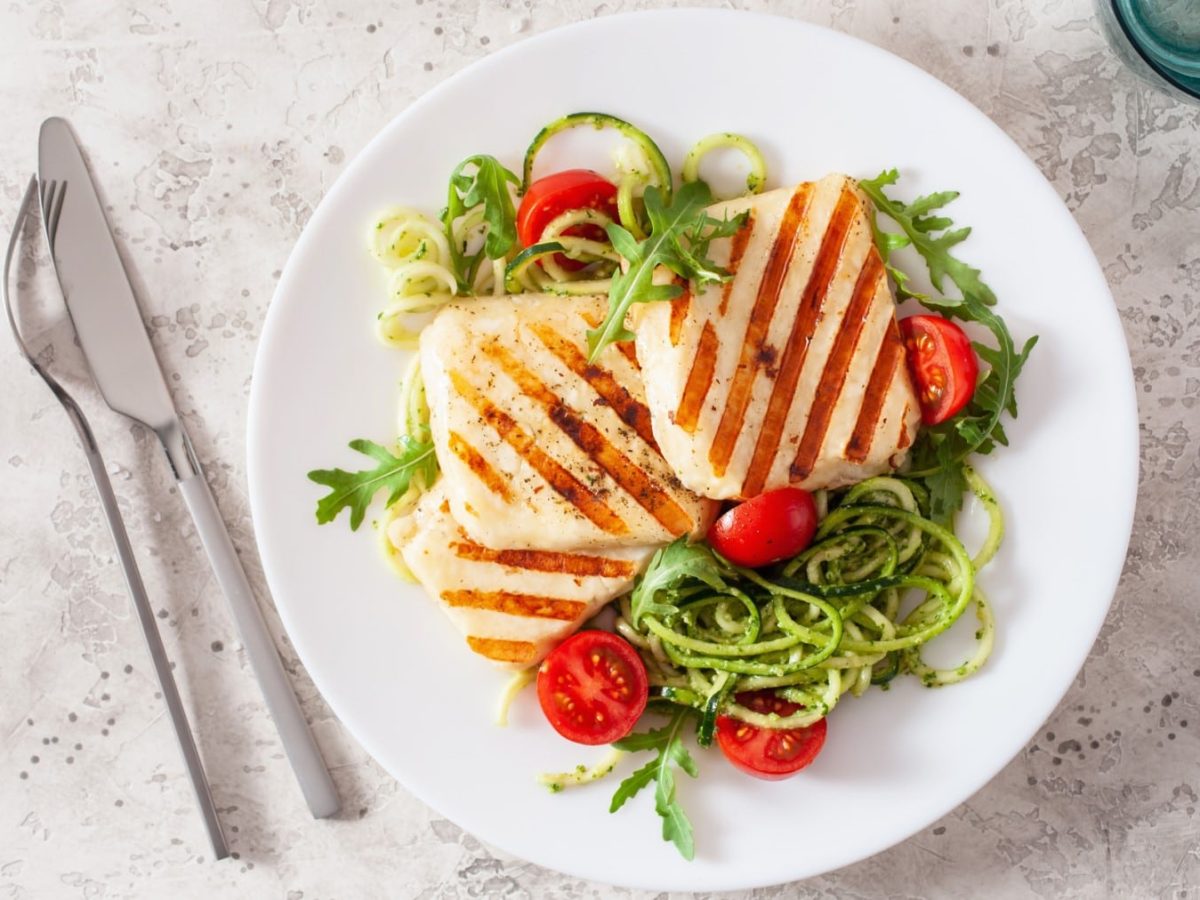 Halloumi auf Zucchinsalat mit grünem Pesto auf einem weißen Teller. Daneben liegt Besteck.