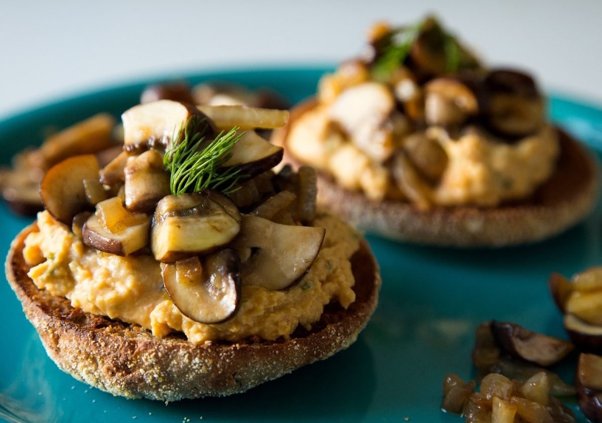 Drei Toasties auf einem blauen Teller mit Hummus und gebratenen Pilzen garniert.