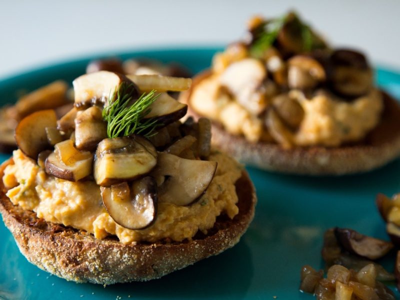 Drei Toasties auf einem blauen Teller mit Hummus und gebratenen Pilzen garniert.