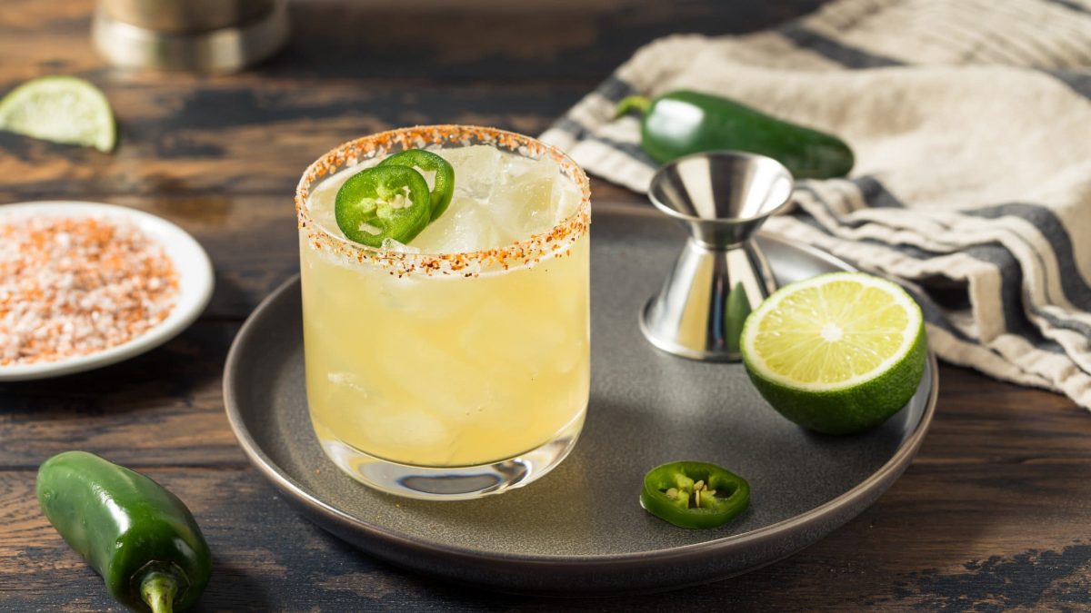 Jalapeno Margarita mit Chili-Salz-Rand, Barmaß, halber Limette und Jalapno-Scheibe auf grauem Teller auf Holztisch. Daneben Jalapeno, Untersetzer mit Chili-Salz und Limette. Rechts Tuch. Frontalansicht.