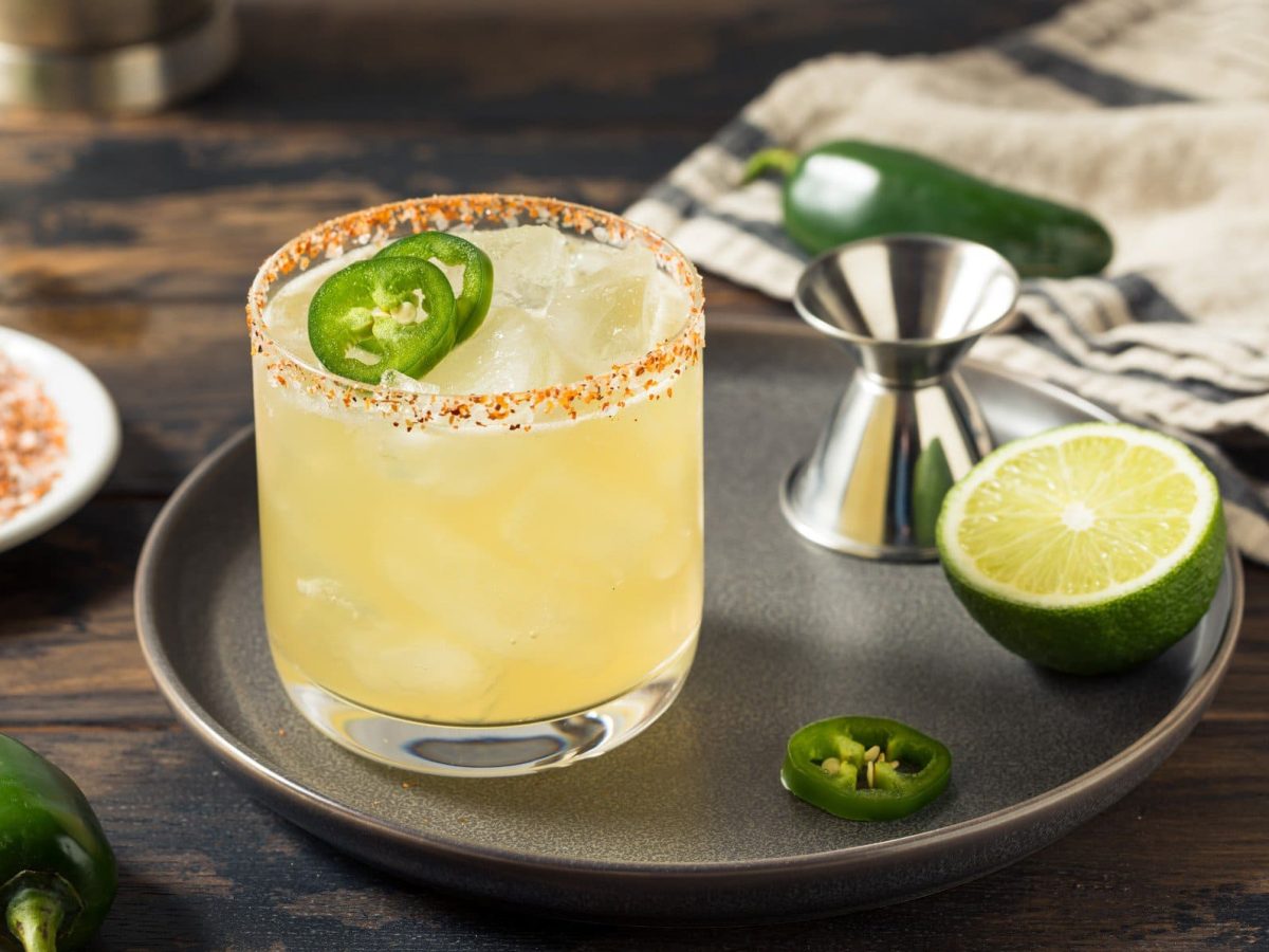 Jalapeno Margarita mit Chili-Salz-Rand, Barmaß, halber Limette und Jalapno-Scheibe auf grauem Teller auf Holztisch. Daneben Jalapeno, Untersetzer mit Chili-Salz und Limette. Rechts Tuch. Frontalansicht.