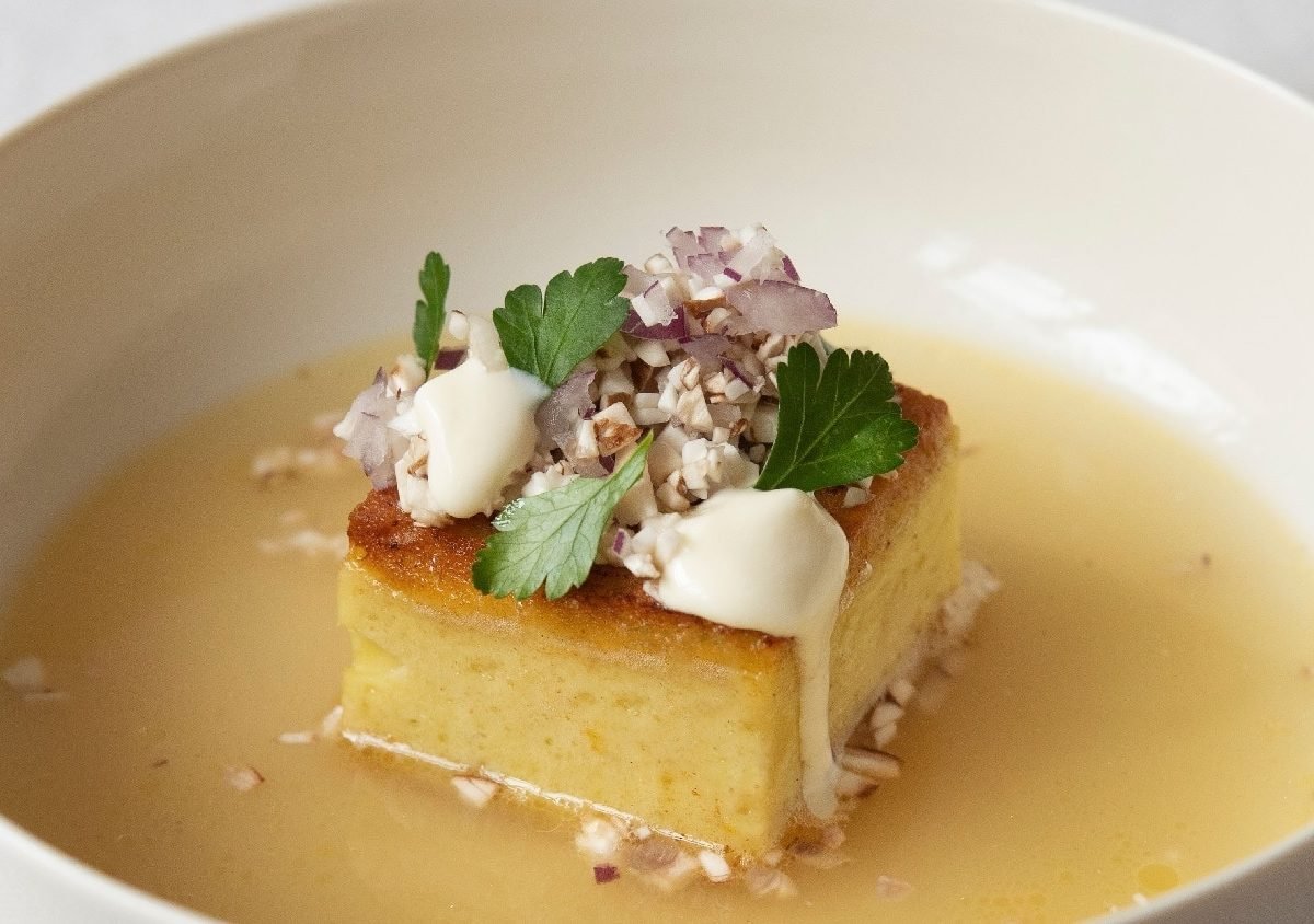 Eine weiße Schale mit goldgelber Brühe und einem viereckigen Kartoffelpudding in der Mitte des Tellers. Auf dem Kartoffelpudding sind Champignons, Creme Double und etwas Petersilie.