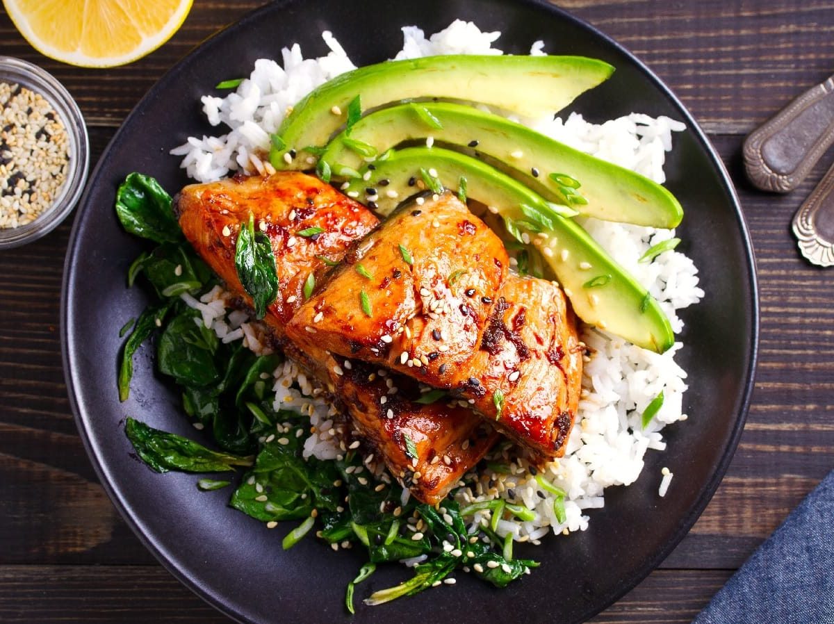 Lachs mit Honig-Soja-Glasur,, Avocado, Spinat und Reis auf einem schwarzen Teller auf einem holzfarbenen Tisch