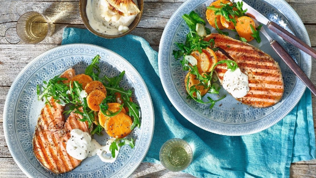 Ein Lachssteak mit Grillmuster auf einem blauen Teller mit Süßkartoffeln und Rucola. Darunter ein Holztisch und eine blaue Decke.