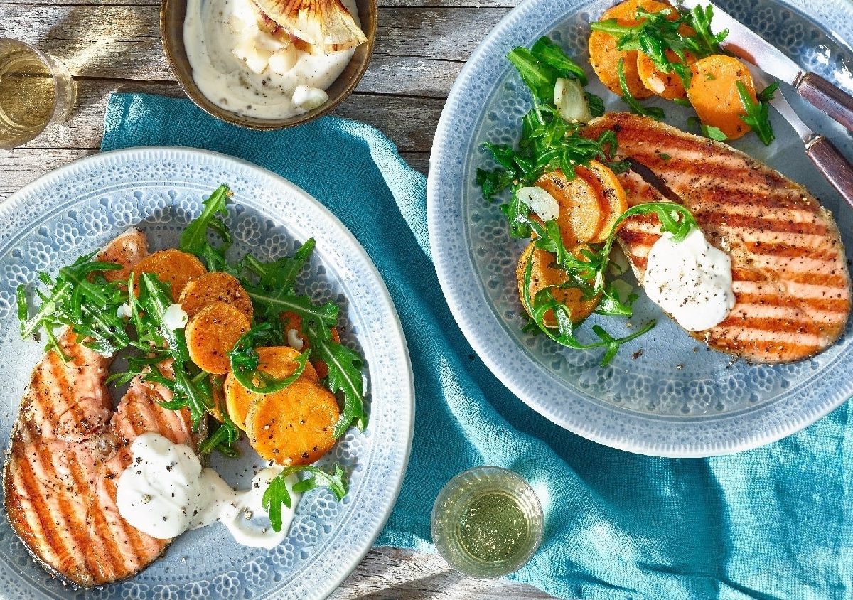 Ein Lachssteak mit Grillmuster auf einem blauen Teller mit Süßkartoffeln und Rucola. Darunter ein Holztisch und eine blaue Decke.