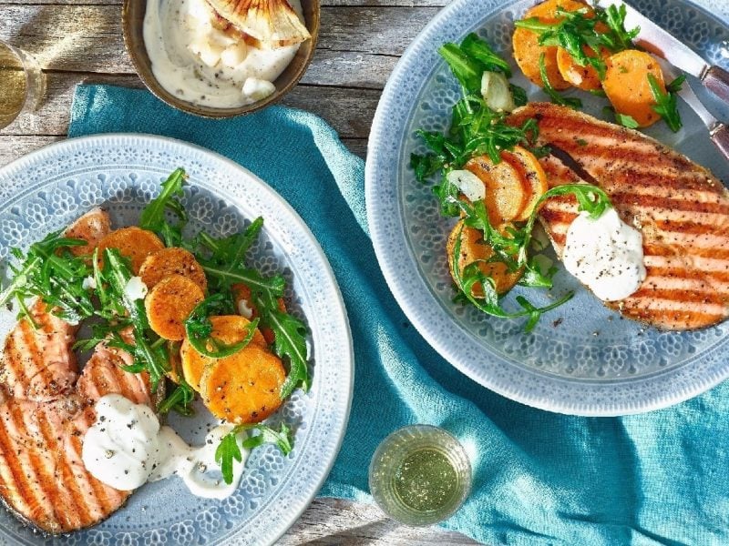 Ein Lachssteak mit Grillmuster auf einem blauen Teller mit Süßkartoffeln und Rucola. Darunter ein Holztisch und eine blaue Decke.