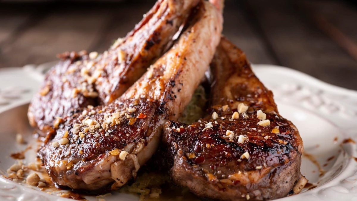 Gegrillte Lammkoteletts mit 5-Pfeffer-Basting und Erdnusscrunch auf weißem Teller und Holzplanken. Frontalansicht.