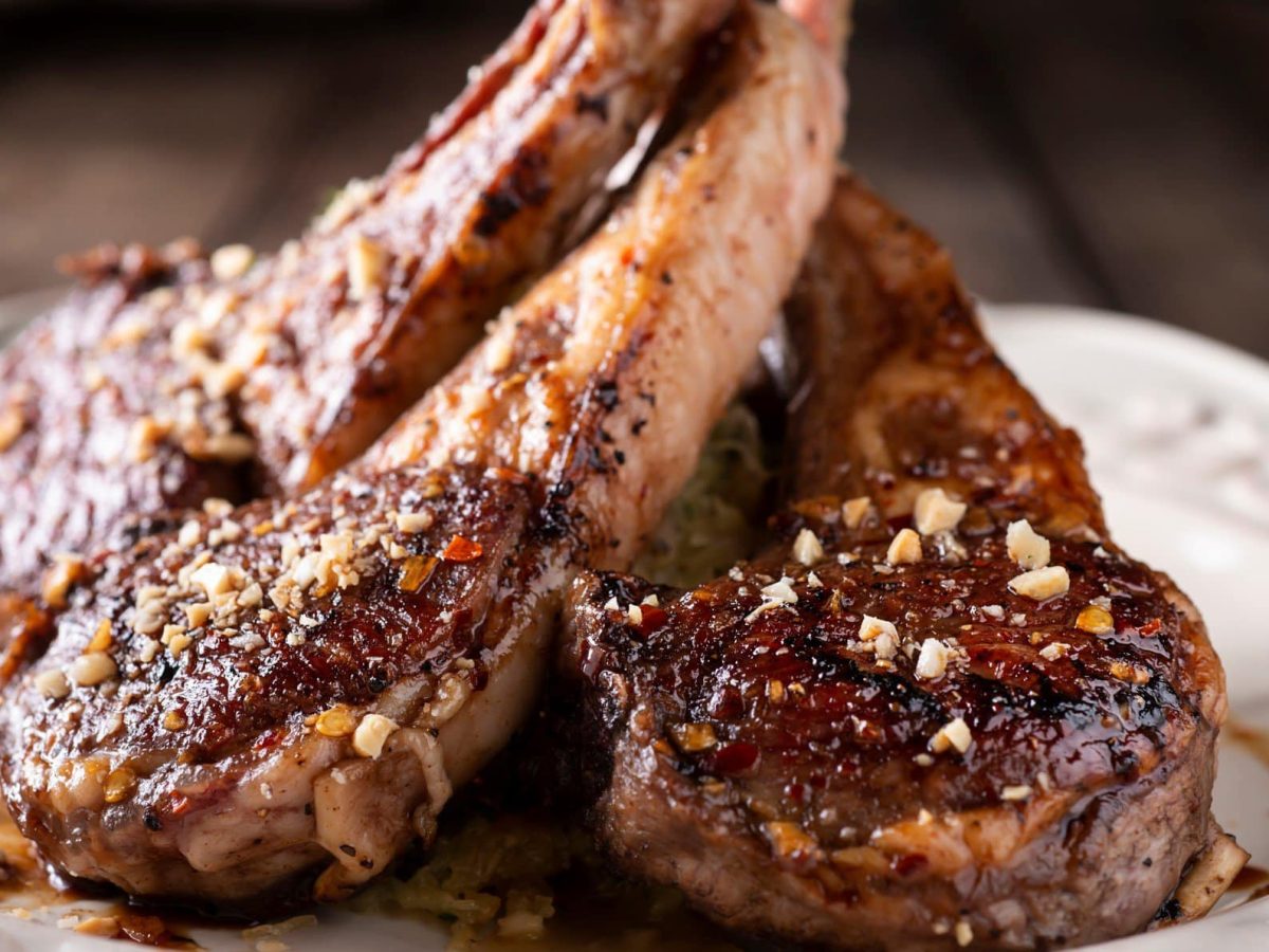 Gegrillte Lammkoteletts mit 5-Pfeffer-Basting und Erdnusscrunch auf weißem Teller und Holzplanken. Frontalansicht.