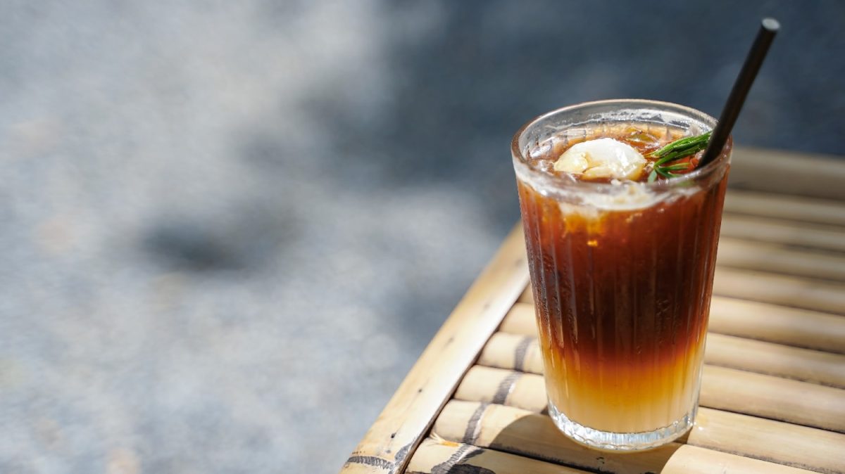 Ein Glas mit dem Litschi-Eistee und einem schwarzen Strohhalm auf einem Holztisch und Betonuntergrund.