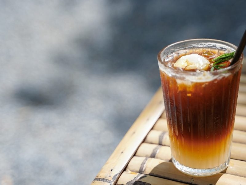 Ein Glas mit dem Litschi-Eistee und einem schwarzen Strohhalm auf einem Holztisch und Betonuntergrund.