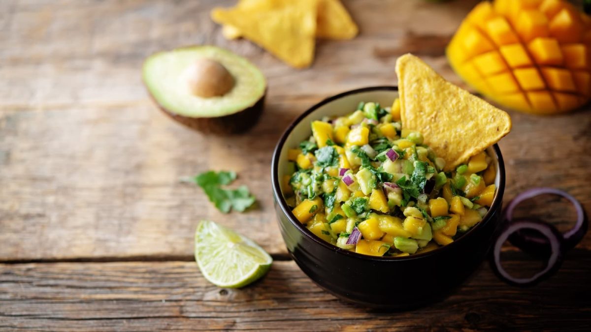 Eine schwarze Schüssel mit Mango Guacamole und einem Tortilla-Chip neben roten Zwiebeln, Limetten, Tortilla-Chips und frischen Mangos.