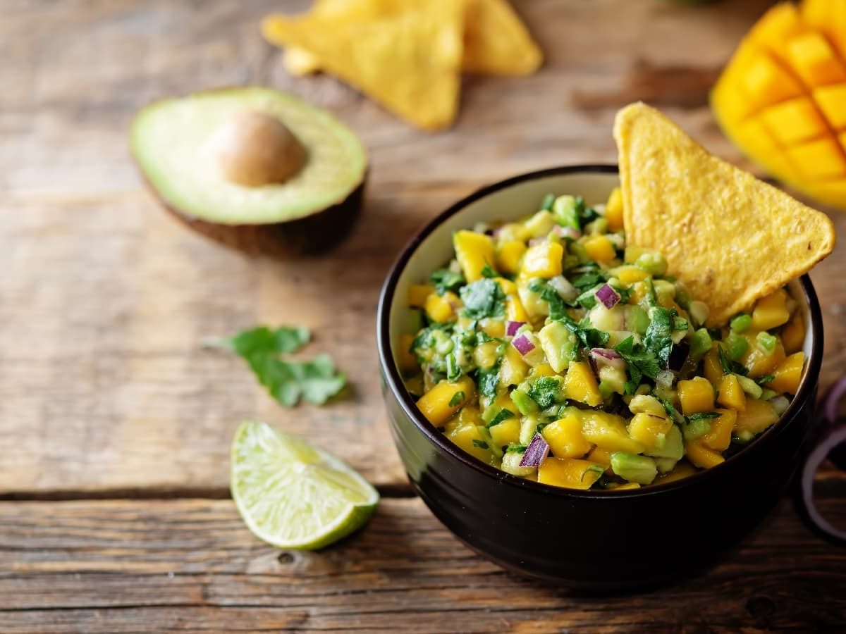 Eine schwarze Schüssel mit Mango Guacamole und einem Tortilla-Chip neben roten Zwiebeln, Limetten, Tortilla-Chips und frischen Mangos.