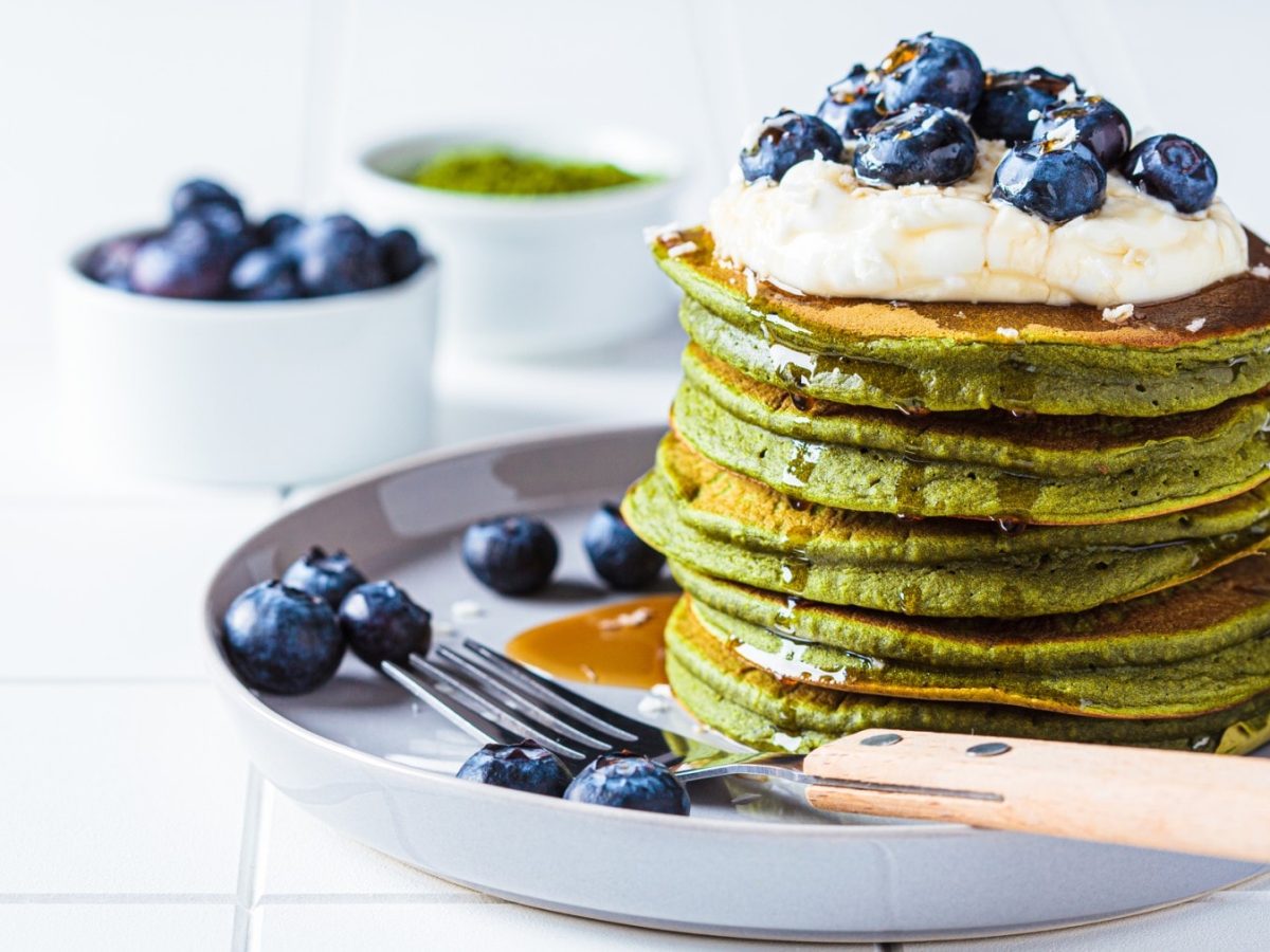 Matcha-Pancakes auf einem grauen Teller. Im Hintergrund zwei Schüsseln mit Heidelbeeren und Matchpulver.