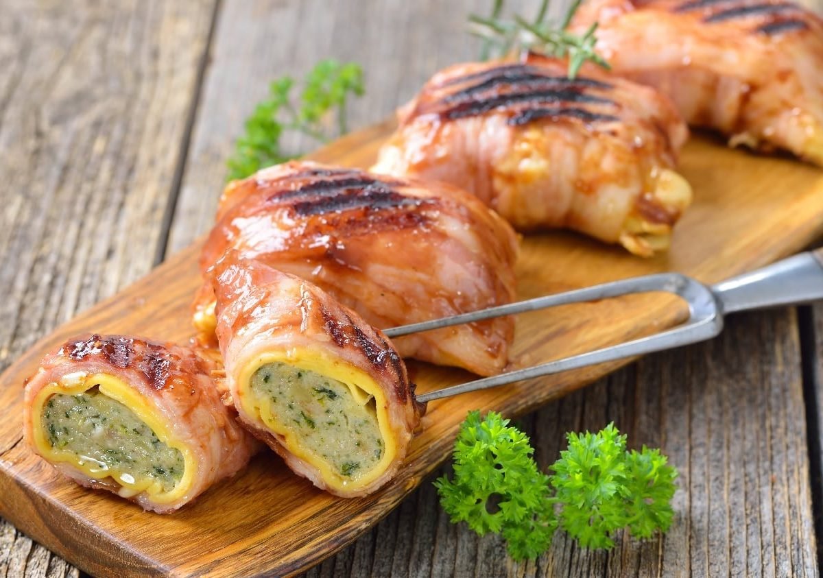 Fünf Maultaschen mit Speck bardiert und gebraten auf einem langen Holzbrett, welches wiederum auf einem Holztisch steht.