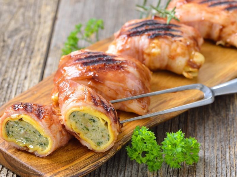 Fünf Maultaschen mit Speck bardiert und gebraten auf einem langen Holzbrett, welches wiederum auf einem Holztisch steht.