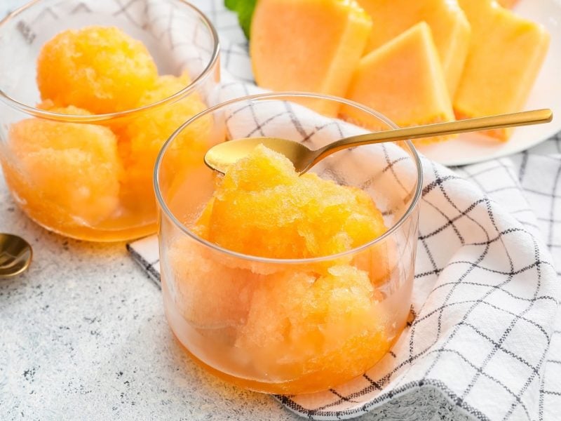 Zwei Gläser Melonensorbet mit Mango und zwei goldenen Teelöffeln, daneben ein Teller Melonenstücke und ein Geschirrtuch.