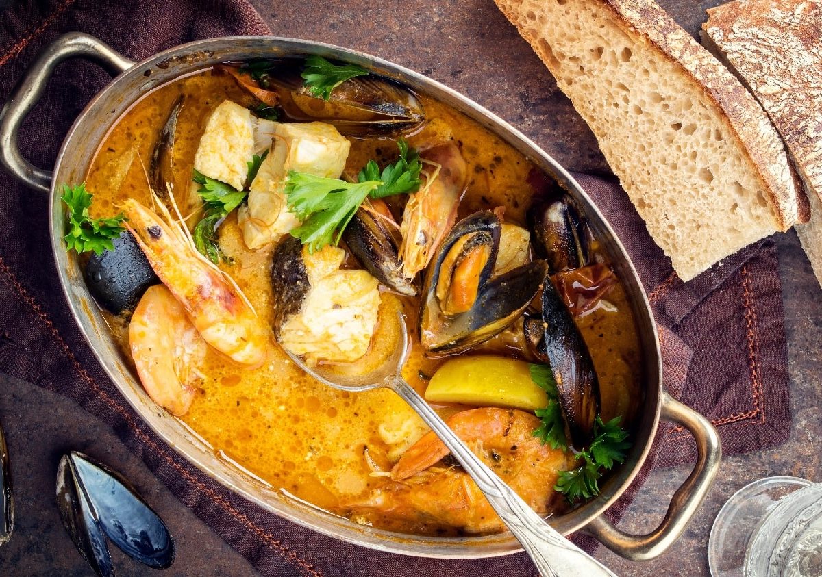 Ein brasilianischer Fischeintopf in einem silberne ovaler Topf. Darunter einige Miesmuscheln und darüber etwas Brot auf einem dunkelbraunen Untergrund.