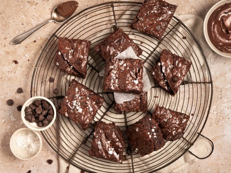 Nutella-Brownies auf Kuchengitter auf hellbraunem Untergrund. Daneben Schälchen mit Meersalz, Schokodrops und Nutella. Oben Teelöffel mit Nutella. Aufnahme aus der Vogelperspektive.