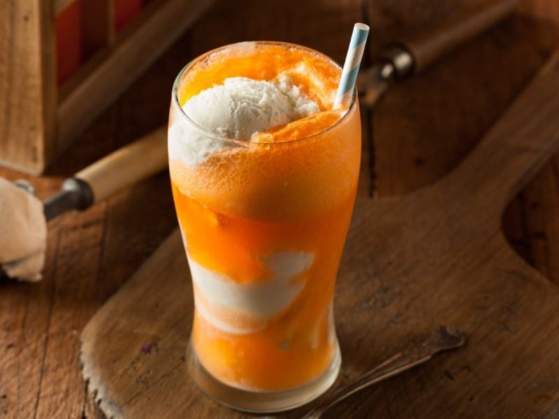 Ein großes Glas mit orange-weißer Schichtung der verschiedenen Orange Cream Float Zutaten. Alles auf einem dunklen Holztisch.