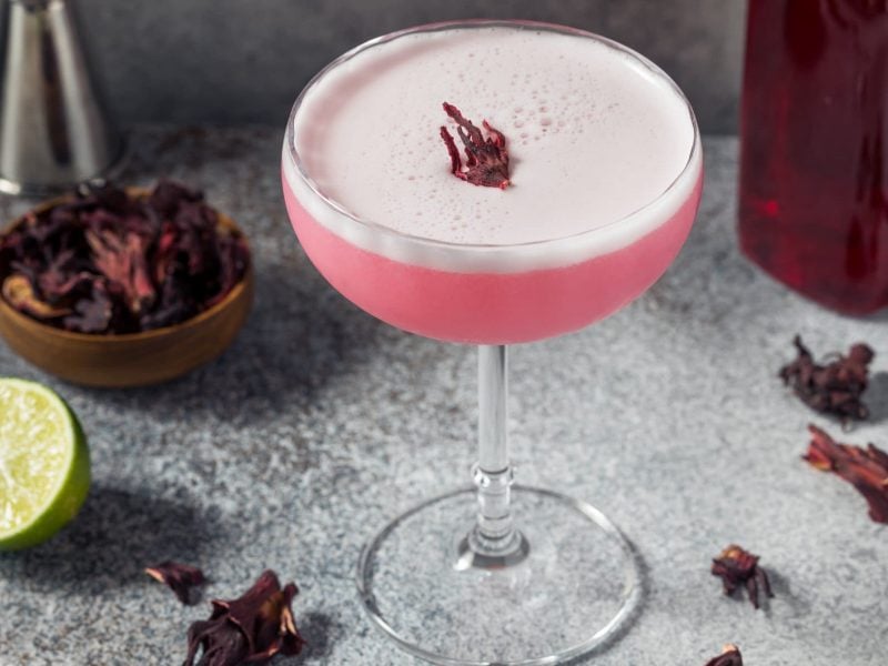 Pink Vodka Sour mit Hibiskus infused Vodka auf grauem Tisch. Daneben Hibiskus-Blüten, Limette und Bar-Maß. Recht eine Flasche mit Hibiskus-Sirup. Frontalansicht.