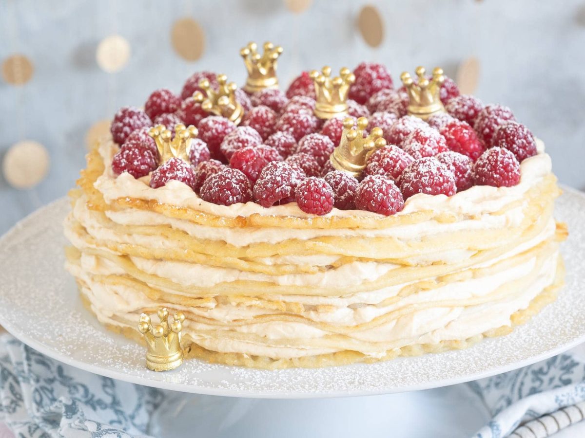 Eine Pfannkuchentorte mit Karamellquark und Himbeeren, dazwischen kleine goldene Krönchen, auf einem Tortenständer. Das Rezept stammt aus dem Kochbuch "Es war einmal".