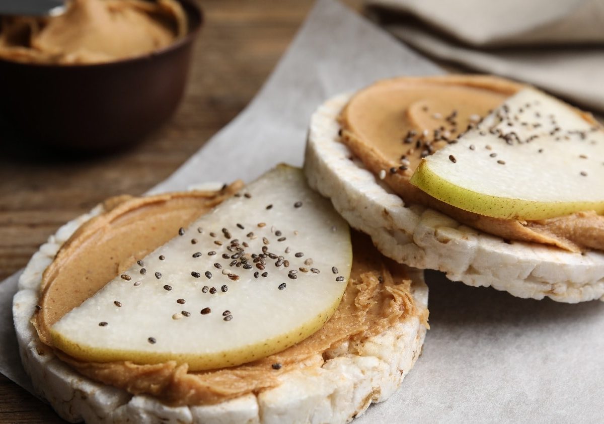 Zwei Reiswaffeln mit hellbrauner Creme auf Pergamentpapier, welches wiederum auf einem Holztisch liegt. Außerdem ist eine Birnenspalte und Chiasamen auf den Reiswaffeln.