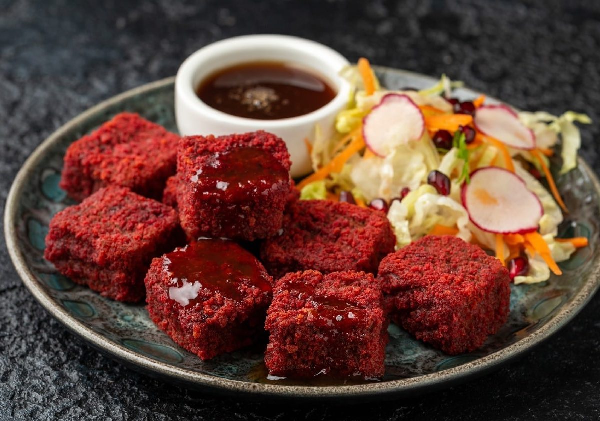 Rote-Bete-Tofu-Würfel auf einem grauen Teller mit Granatapfelsauce und etwas gemischtem Salat. Alles auf einem dunklen Untergrund.