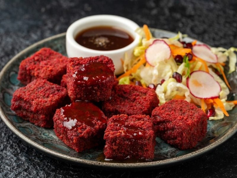 Rote-Bete-Tofu-Würfel auf einem grauen Teller mit Granatapfelsauce und etwas gemischtem Salat. Alles auf einem dunklen Untergrund.