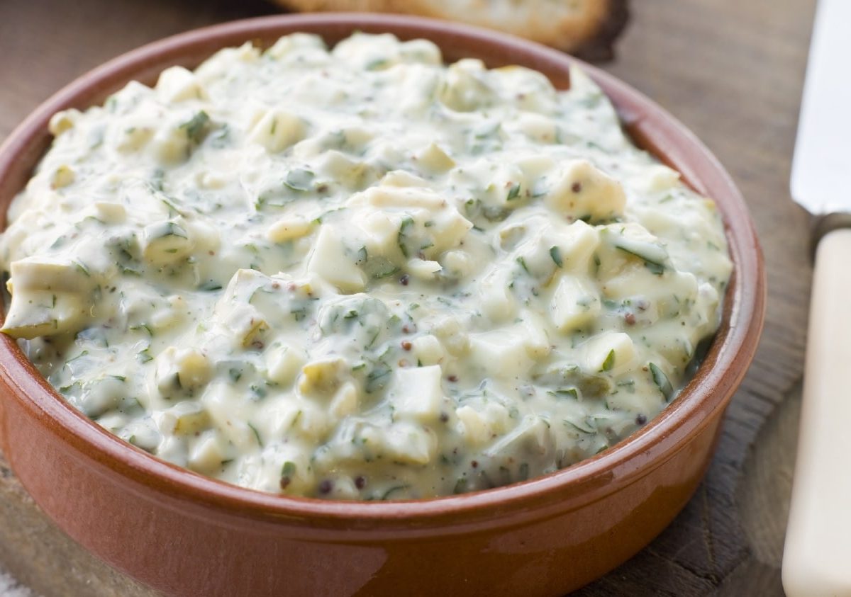 Eine braune Schale mit der Sauce Gribiche auf einem Holztisch. Im Vordergrund ein Messer und im Hintergrund ein paar geröstete Scheiben Weißbrot.