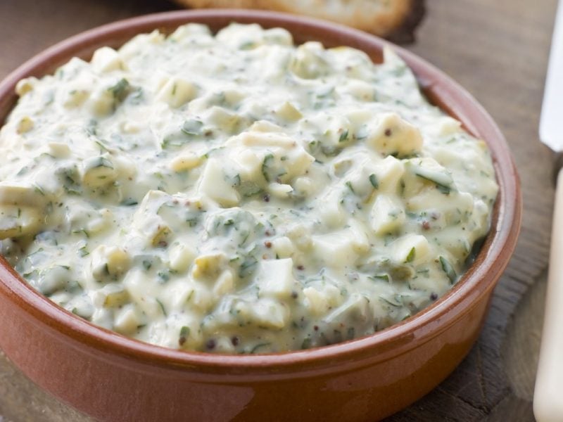 Eine braune Schale mit der Sauce Gribiche auf einem Holztisch. Im Vordergrund ein Messer und im Hintergrund ein paar geröstete Scheiben Weißbrot.