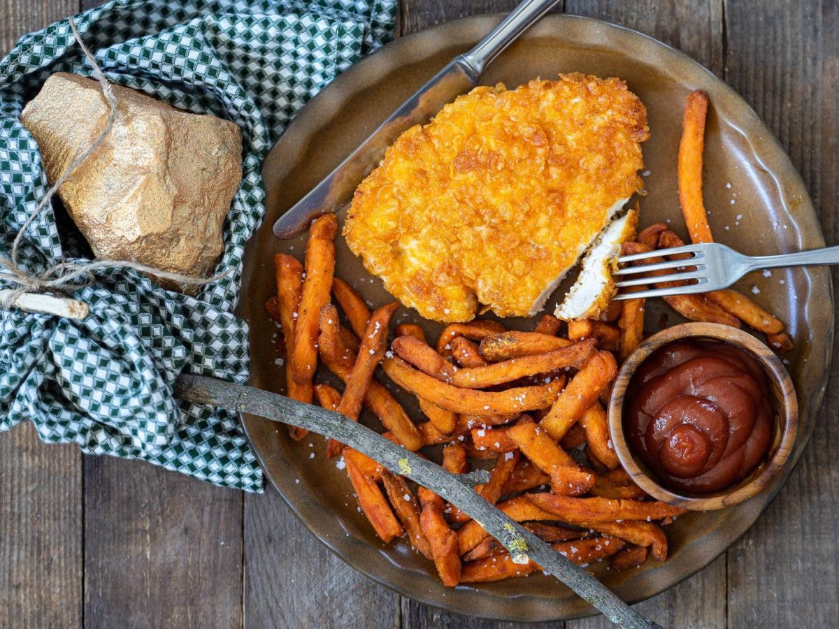 Ein Schnitzel mit Cornflakes-Panade, Süßkartoffeln und einer Schale Ketchup auf einem Teller in der Draufsicht, daneben ein Tuch mit einem kleinen Goldklumpen.