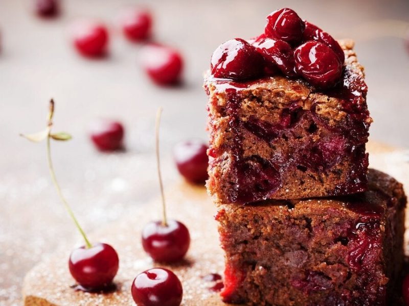 Zwei Schoko-Brownies mit Kirschen stehen auf einem Brettchen.