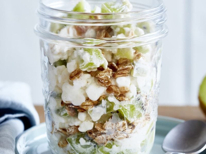 Schicht-Flocken aus dem Glas mit Kiwi stehen auf einer Untertasse. Daneben ein Löffel und eine aufgeschnittene Kiwi.