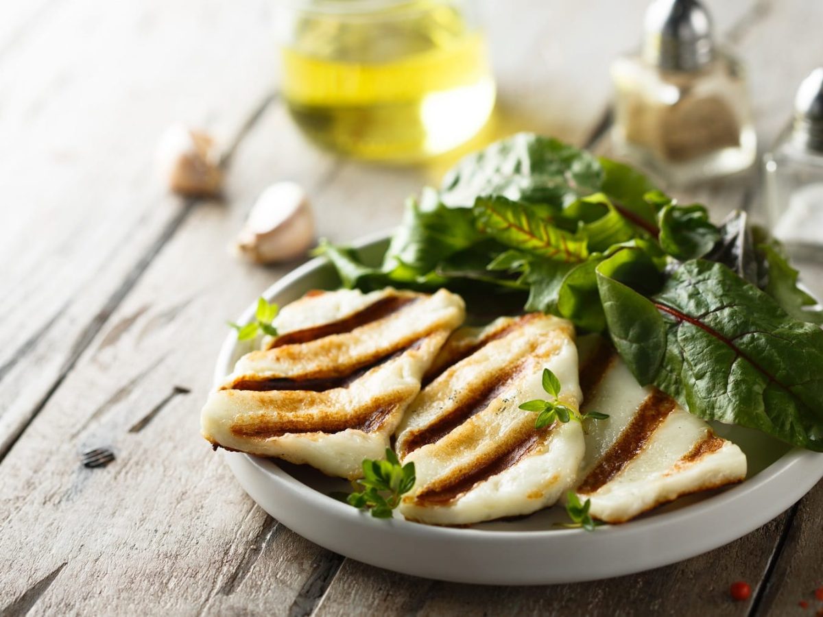 Ein Teller mit ein paar Scheiben selbst gemachtem veganem Halloumi und grünem Salat, dahinter unscharf Öl, Gewürzstreuer und Knoblauchzehen.