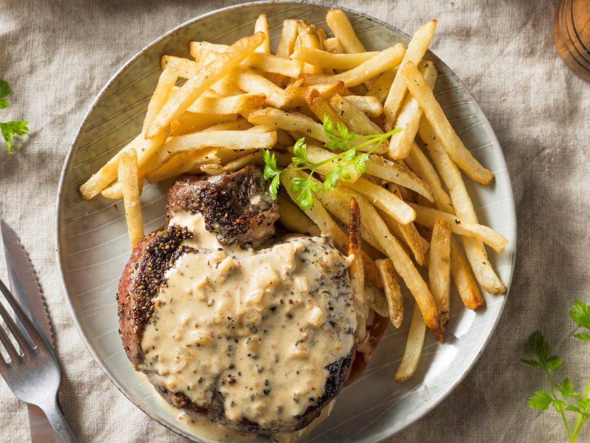 Ein heller Teller mit Pommes und dem Steak au poivre auf einem beigen Tuch. Dazu ein paar stängel Petersilie die auf dem Tisch verteilt sind.