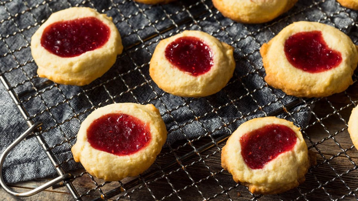 Einige Thumbprint Cookies auf einem Metallgitter und einem Holztisch. Oben links im Bild ist außerdem ein grauer Stoffuntersetzer zu sehen.