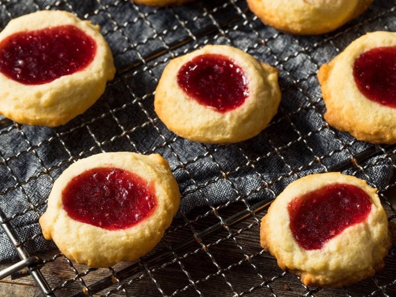 Einige Thumbprint Cookies auf einem Metallgitter und einem Holztisch. Oben links im Bild ist außerdem ein grauer Stoffuntersetzer zu sehen.