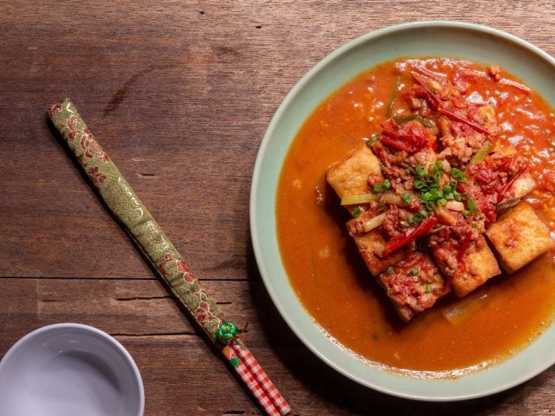 Eine Schüssel Tofu in Tomatensauce neben einem Paar Stäbcgen