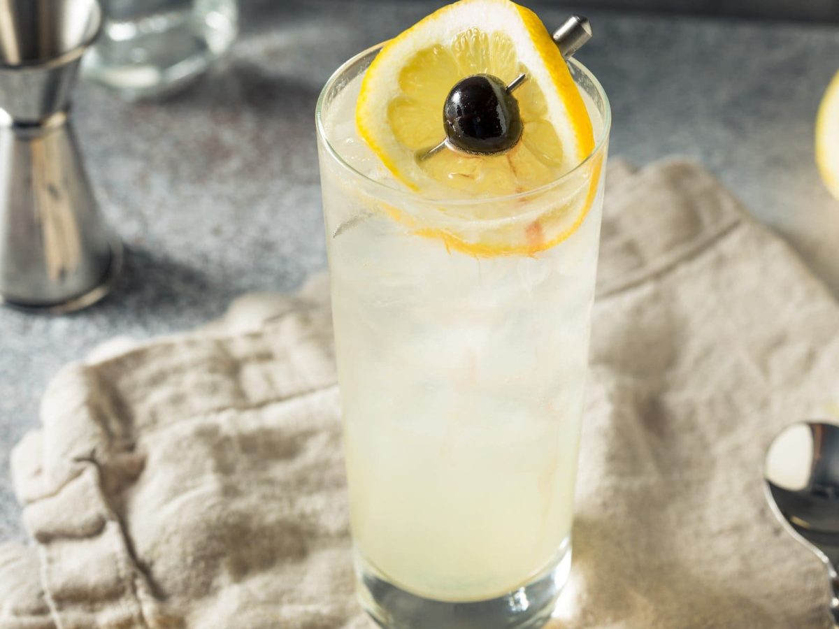 Tom Collins Gin-Cocktail auf beigem Tuch auf grauem Tisch mit Zitronenscheibe und Kirsche auf Metallspieß. Daneben Barmaß. Im Hintergrund eine halbe Zitrone. Frontalansicht.