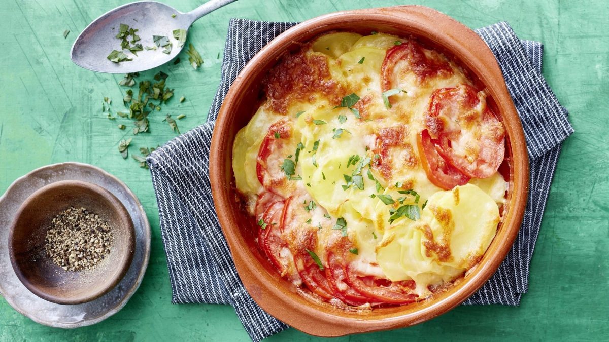 Tomaten-Kartoffel-Gratin in einer Auflaufform auf einem Küchentuch. Daneben Besteck und verschiedene Zutaten.