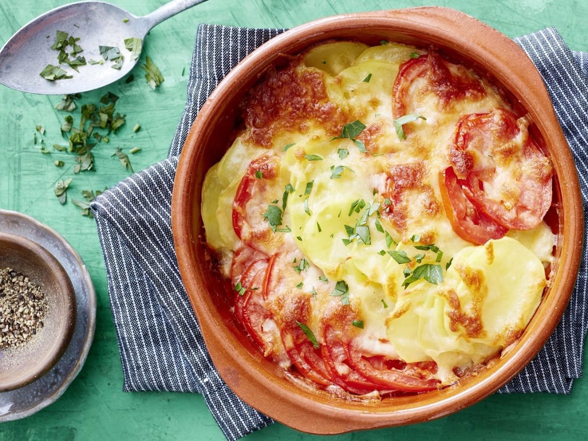 Tomaten-Kartoffel-Gratin in einer Auflaufform auf einem Küchentuch. Daneben Besteck und verschiedene Zutaten.