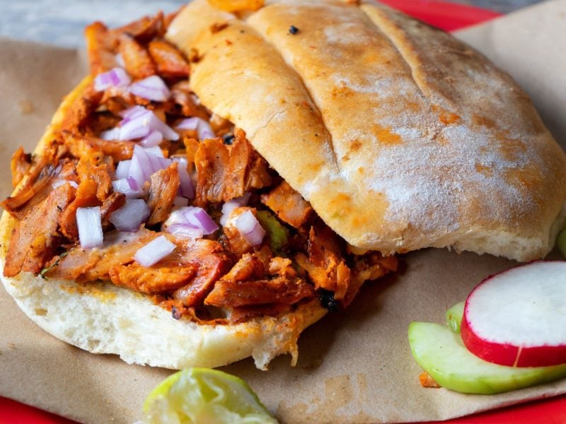 Tortas al Pastor auf braunem Papier auf rotem Teller auf blauem Untergrund. Daneben eine Scheibe Radieschen und Limette. Draufsicht.