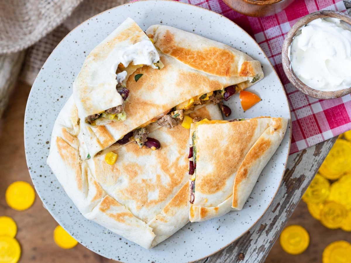 Tortilla-Crunchwraps mit Hackfleisch auf einem Teller in der Draufsicht, daneben eine Schale mit weißem Dip und ein paar Goldtaler.