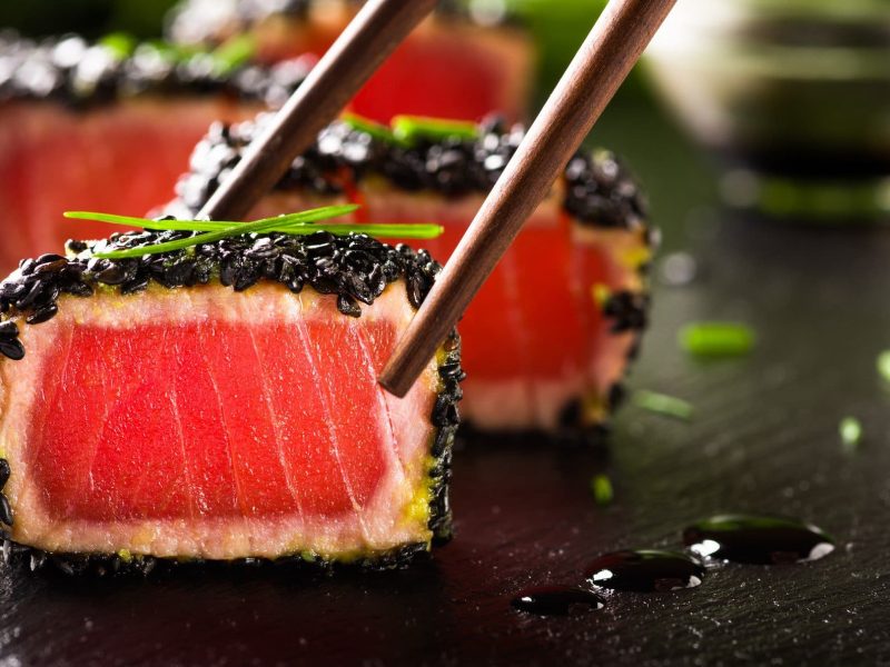 Tunatataki mit schwarzem Sesam und Miso-Creme auf schwarzer Granitplatte. Im Hintergrund Sauce in Glasschüssel. Zwei Stäbchen von oben ins Bild ragend. Frontalansicht.